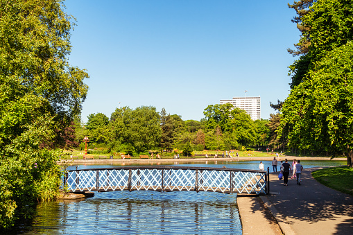 Glasgow, Scotland - A bridge over the pond in Victoria Park in Glasgow's West End, with people walking around the pond's edge on a sunny day.