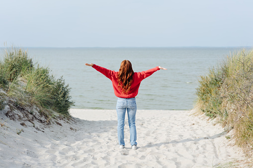 Young woman rejoicing at the warmth of the fall or autumn sun standing on a sandy footpath overlooking a tropical beach with outstretched arms