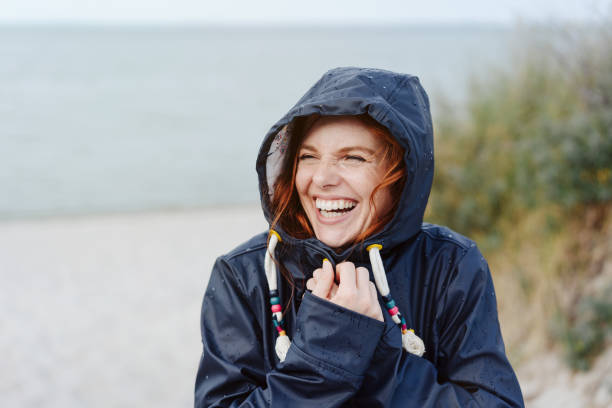 Laughing happy woman embracing the cold weather Laughing happy young woman embracing the cold autumn weather snuggling into her warm anorak with a beaming vivacious smile as she strolls along a beach raining outside stock pictures, royalty-free photos & images