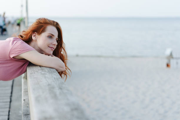 Attractive serene young woman standing leaning on the parapet of a wooden pier overlooking the beach watching below with a quiet smile and dreamy expression