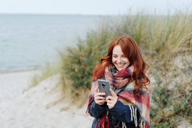 Happy young woman enjoying the beach in winter Happy young woman enjoying the beach in winter standing on the dunes above the sea reading a text message on her mobile phone with a smile baltic sea people stock pictures, royalty-free photos & images