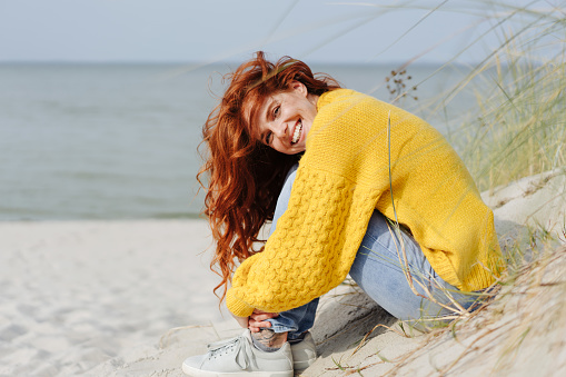 Smiling vivacious young woman relaxing on a tropical beach sitting on the sand dunes overlooking the sea enjoying the autumn sunshine