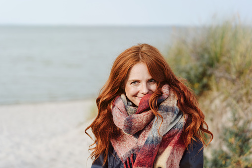 Happy young woman with long red hair wearing a warm woolly scarf and anorak walking on a beach in winter smiling at the camera