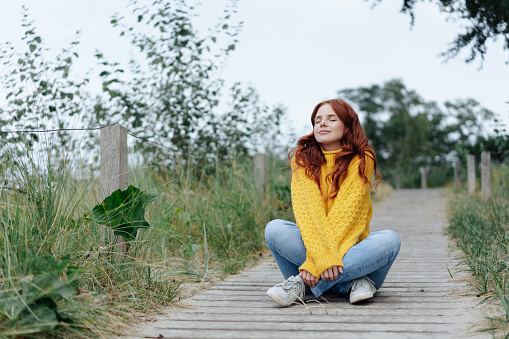 Young woman relaxing cross-legged on a coastal boardwalk with closed eyes and a blissful smile of contentment on a cold autumn day