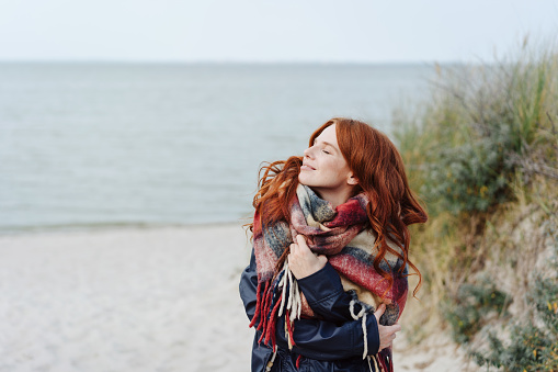 Active healthy young woman enjoying a sandy tropical winter beach standing in a warm anorak and scarf looking up into the sun with a blissful smile and closed eyes