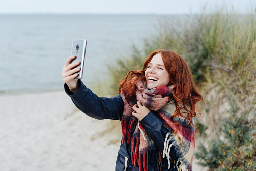 Laughing young woman wearing a warm winter scarf taking a selfie on a mobile phone as she enjoys a day of autumn sunshine on a tropical beach and dunes