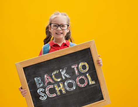 Back to school and happy time! Cute industrious child is holding blackboard on color paper wall background. Kid with backpack. Girl ready to study.