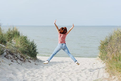 Girl in pink t-shirt and blue jeans jumping on the beach with her legs and arms spread in the air, enjoying the sea view, shot from her back