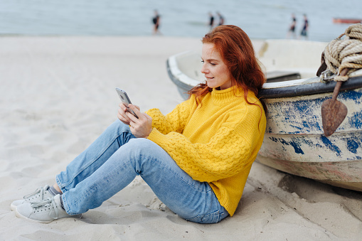 Young woman relaxing on a beach with her mobile phone sitting on the sand leaning against an old wooden rowboat
