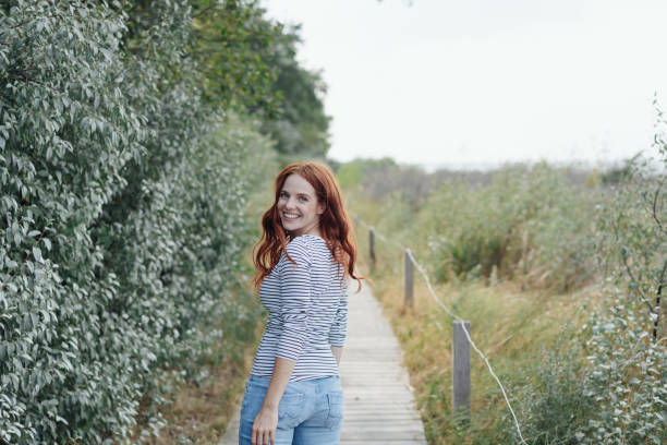 Pretty young redhead woman turning to the camera with a vivacious happy friendly smile as she looks back over her shoulder while walking along a boardwalk
