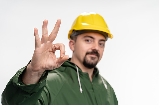 Positive young engineer with green raincoat and hardhat  showing OK sign on white background, close-up. Photo is taken in studio environment with Sony A7III camera