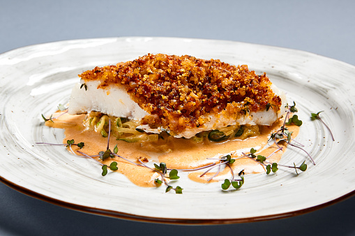 Baked cod fillet with sauce. Restaurant food portion side view. Fish with batter garnish served on white craft plate. Seafood sprinkled with breadcrumbs meal course. Luxury gourmet dish serving