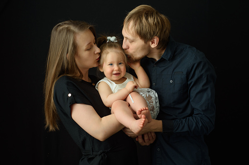 Father and mother kiss their little one year old daughter. Shot on a black background.