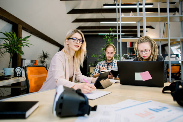 istock Group of young creative designers, business people, gathered together to discuss idea, develop startup. Two pretty girls and handsome guy working at the office table using electronics gadgets 1251517317