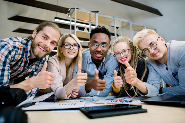 istock Group of skilled five multiethnic designers, architects, freelancers, working together on project, leaning on the table, smiling to camera and showing thumbs up. Attractive hipster business people 1251518821