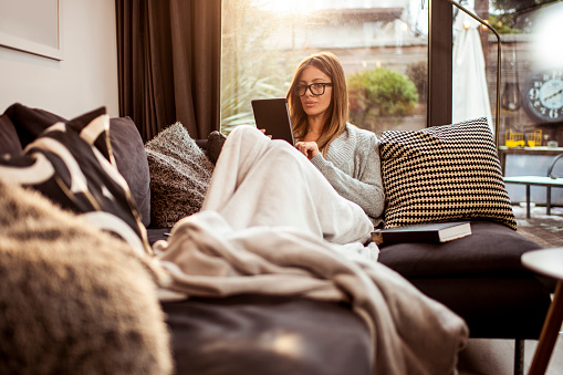 A contemporary woman enjoying the coziness of her home by reading a book on a digital tablet.