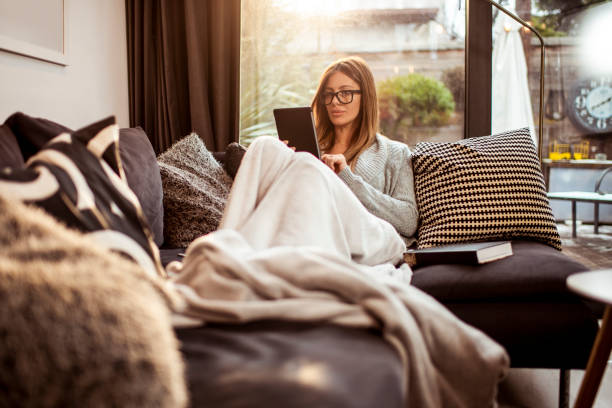 A single woman reading an e-book on her digital tablet while lying on her sofa at home A contemporary woman enjoying the coziness of her home by reading a book on a digital tablet. sofa bed stock pictures, royalty-free photos & images