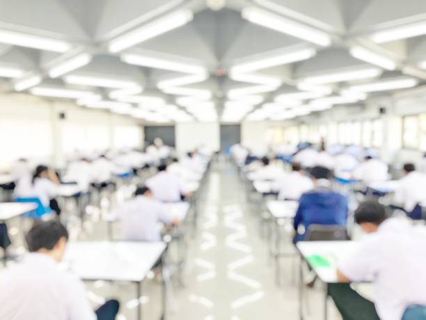 istock Blur abstract background of examination room with undergraduate students inside. Blurred view of student doing final test in exam hall. Blurry view of study chairs in classroom of university or campus 1251528360