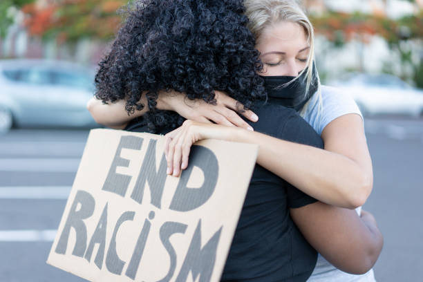 oung donna africana che abbraccia una donna bianca del nord dopo una protesta - donna del nord con fine razzismo bannner nelle sue mani - concetto di nessun razzismo - anti racism foto e immagini stock