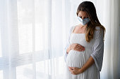 Pregnant woman standing by the big window with face medical mask on