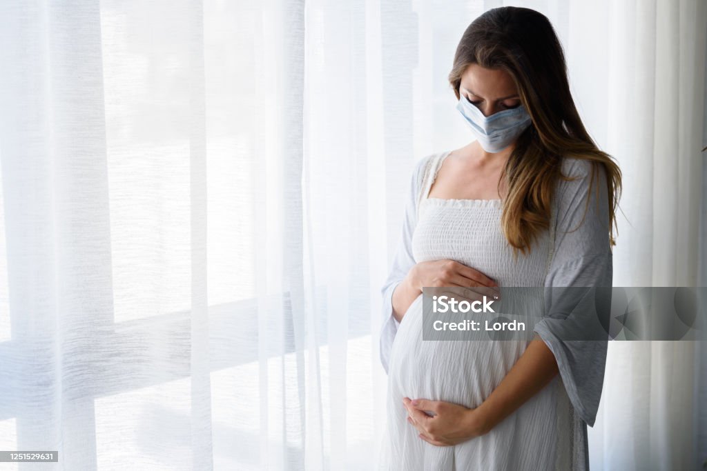 Pregnant woman standing by the big window with face medical mask on Pregnant woman standing by the big window with face medical mask on. Worries about child birth during pandemic. Pregnant Stock Photo
