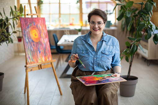 Portrait of happy female artist at atelier, sitting, looking at camera and smiling
