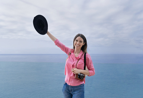 Young traveler woman laughing with a vintage camera and hat - ocean view in the background - Focus on her face