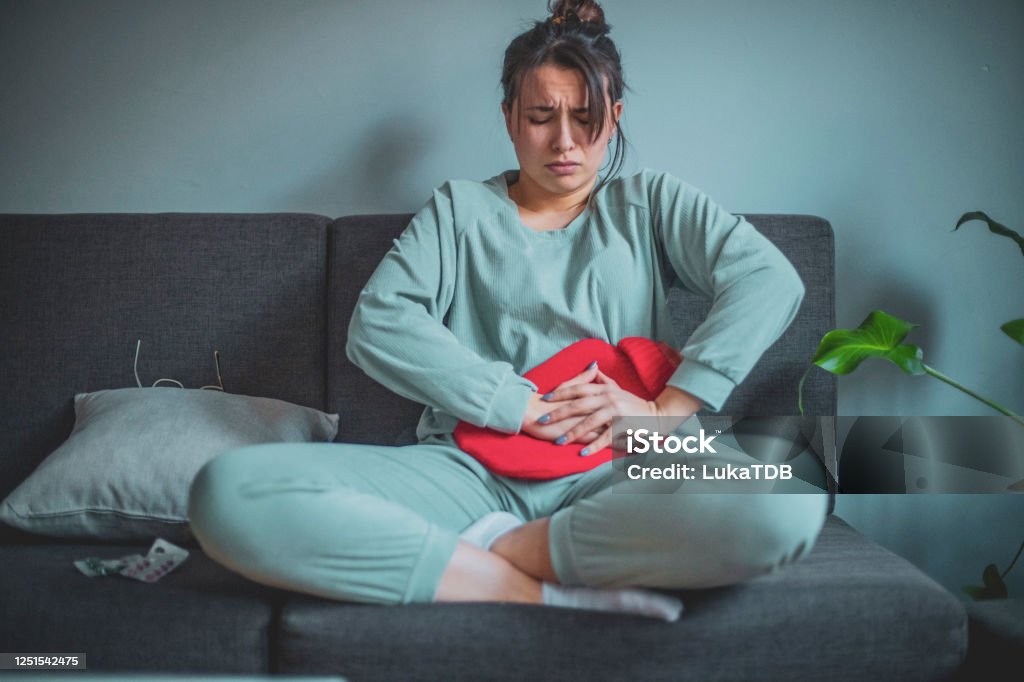 Feeling unwell A young woman is in her home, wearing her pajamas. She's expressing pain on her face as she holds a heating pad pressed onto her stomach. Menstruation Stock Photo