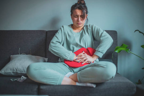 Feeling unwell A young woman is in her home, wearing her pajamas. She's expressing pain on her face as she holds a heating pad pressed onto her stomach. pms photos stock pictures, royalty-free photos & images