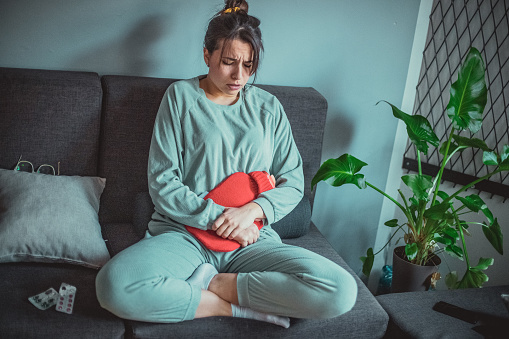A young Caucasian woman is feeling under the weather and expressing her pain. She's in her pajamas in her living room and  she's treating abdominal spasms and cramps with a thermal water bag.