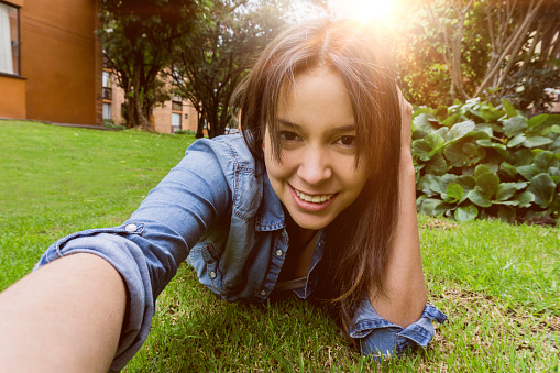 Pretty young woman taking a selfie at the park while lying down on her tummy facing camera smiling
