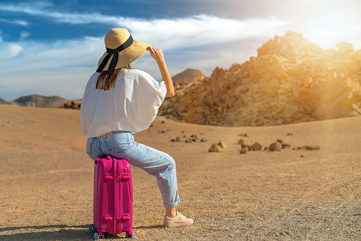 Girl in a white shirt and hat sit on a pink suitcase on a volcanic sandy scenery. Teide volcano, Tenerife, Spain