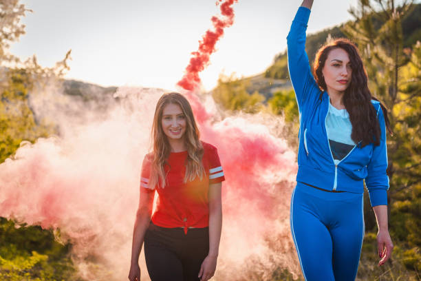 Young woman wearing a smoking torch above her head. The other girl is following her and looking at camera with a smile There is red smoke behind them.