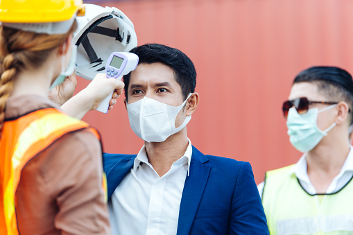 Female worker scanning fever temperature with digital thermometer construction site staff wearing hygiene face mask protect from Coronavirus or COVID-19. New Normal working life adaptation in 2020