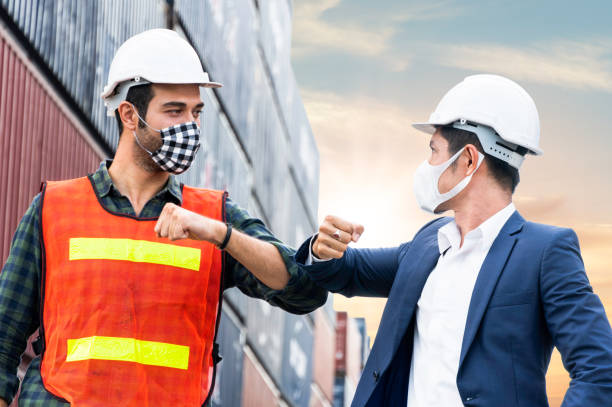 コロナウイルスまたはcovid-19が広がるのを防ぐために新法適応で衛生フェイスマスク肘のバンプ挨拶を身に着けている建設現場の労働者と職員。倉庫での社会的距離の作業スタイル - helmet stack safety hardhat ストックフォトと画像