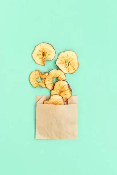 Photo of Healthy snack. Homemade dehydrated fruit chips of apple on mint colored background. Dry apple in paper package. Diet food. Top view and copy space.