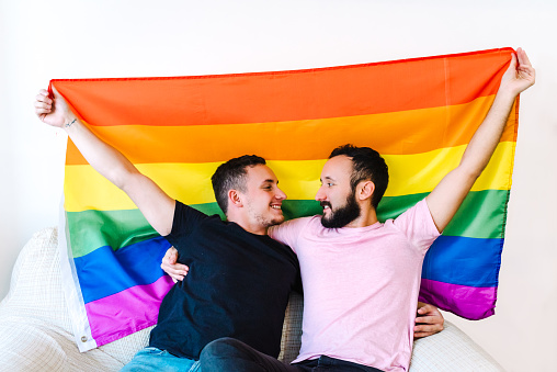 Stock photo of two caucasian homosexual men holding each other and holding up an LGBTQ flag. They have their eyes closed.