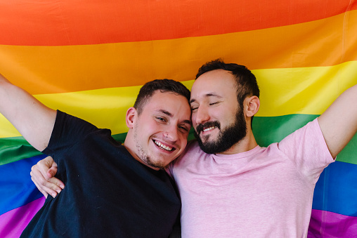 Stock photo of two caucasian homosexual men holding each other and holding up an LGBTQ flag. They have their eyes closed.