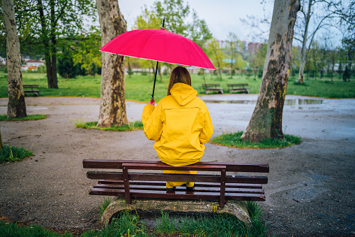 Alone woman sitting on a bench outdoors in the rain, holding red umbrella