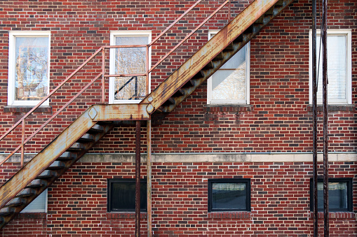 set of back alley rusted fire escape stairs in front of white windows on a red brick building