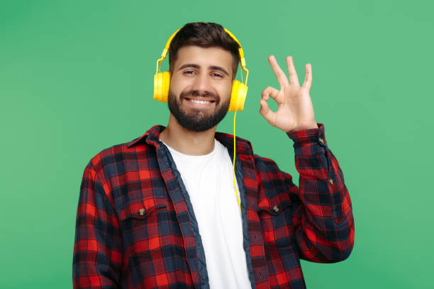 ヘッドフォンを着用し、緑の背景の上にokジェスチャーを示すチェック柄のシャツを着たポジティブなひげを生やしたヒップスターの若者。 - free standing audio ストックフォトと画像