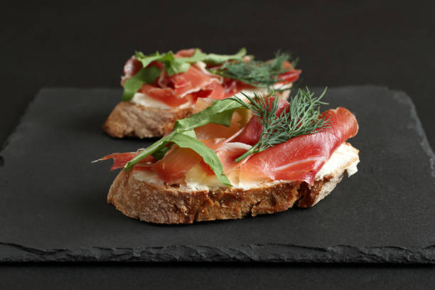 buckwheat toasts with goat cheese, prosciutto and arugula lying on a black stone board on a black table. - appetizer bruschetta meal lunch imagens e fotografias de stock