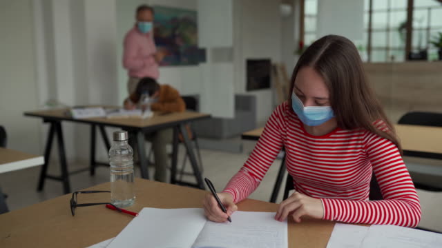 コロナウイルスパンデミック中の教室で、教室に座って、書き込みとカメラを見て保護フェイスマスクを持つ女子高生