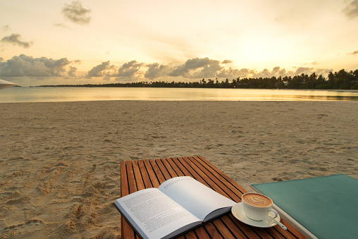 Open book and cup of coffee on the wooden table with sunset beach background