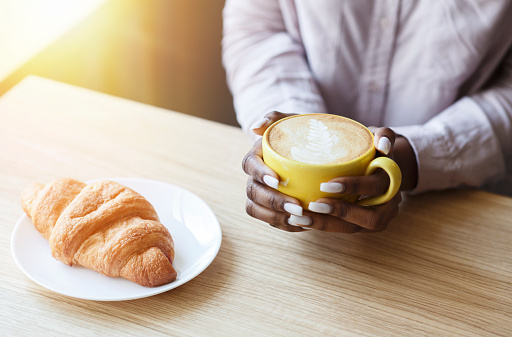Peaceful morning. Unrecognizable African American girl having coffee with croissant at table in cozy cafe, closeup view