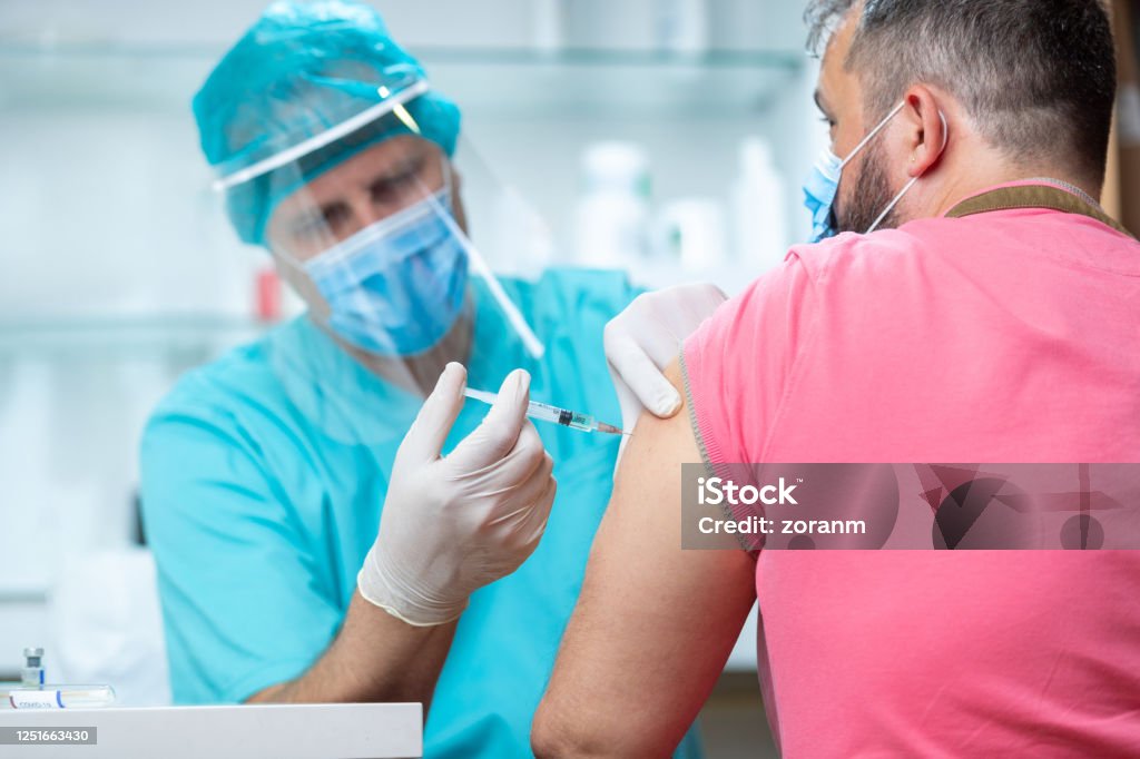 Doctor wearing protective workwear injecting vaccine into patient's arm Doctor wearing protective visor and surgical gloves injecting COVID-19 vaccine into patient's arm Vaccination Stock Photo
