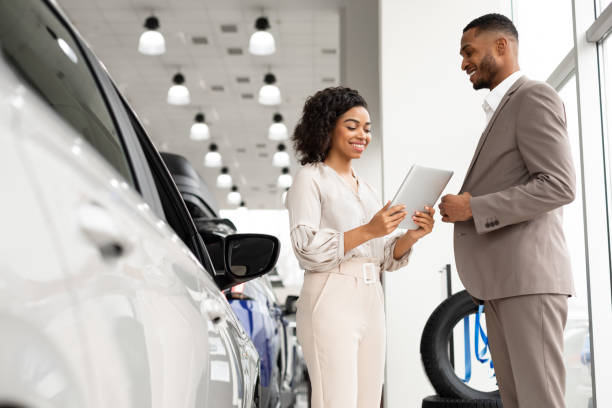 Seller Woman Showing Auto To African Businessman In Dealership Showroom Car Sales. Automobile Seller Woman Showing Auto To African Businessman Standing In Dealership Showroom. Free Space car dealership stock pictures, royalty-free photos & images