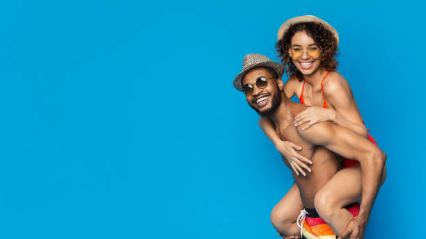 Summer Time. Joyful balck couple in swimwear having fun over blue background Summer Time. Cheerful black girl piggybacking her happy boyfriend, joyful couple in swimwear having fun on blue background, panorama with free space black woman bathing suit stock pictures, royalty-free photos & images