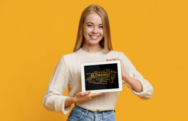 istock Smiling blonde girl showing tablet computer with modern blogging terms on screen, orange background. Collage 1251669885
