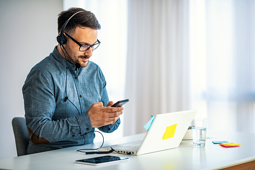 Young happy businessman smiling while reading his smartphone. Portrait of smiling business man reading message with smartphone in office. Man working at his desk at office.